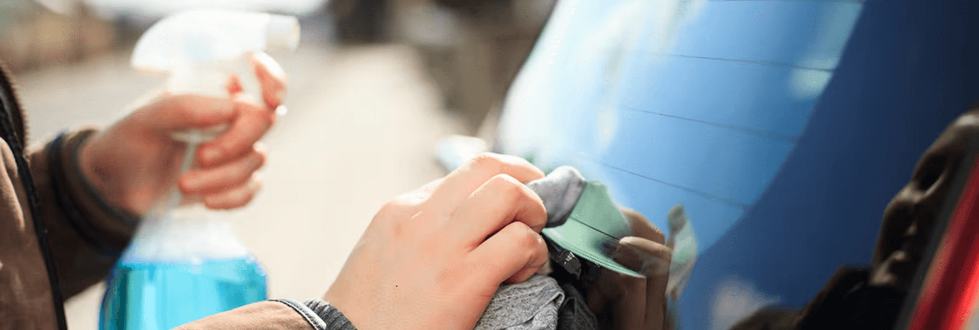 Back windshield of car getting cleaned by person holding a spray bottle containing blue liquid.